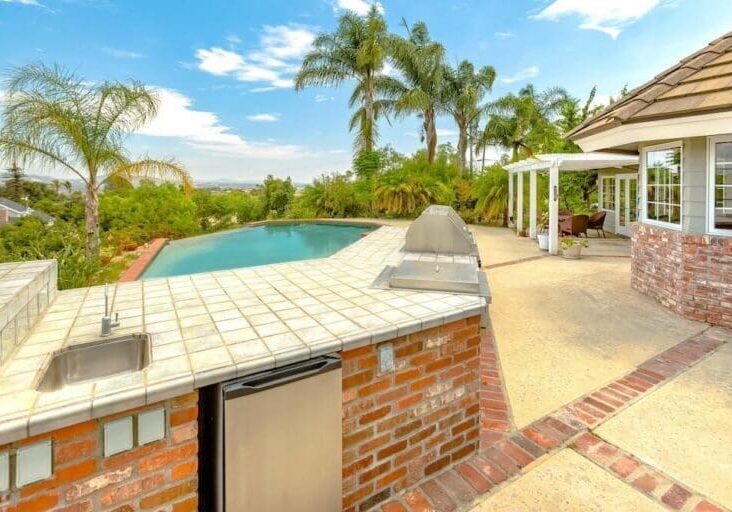 Brick outdoor kitchen with tile countertop overlooking a backyard pool and palm trees