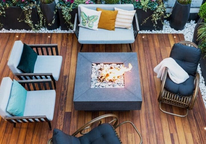 Aerial view of a modern wood deck with square concrete fire pit, cushioned lounge seating, and white pebble garden borders