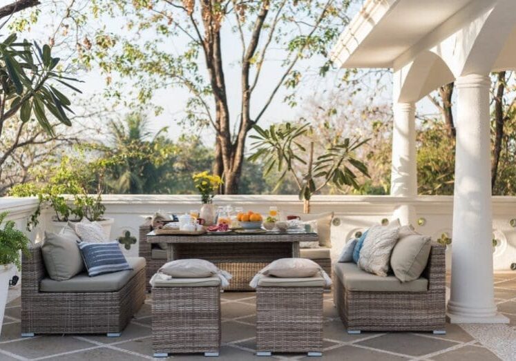 Elegant covered terrace with wicker lounge furniture, decorative pillows, and a fruit-filled coffee table overlooking lush trees