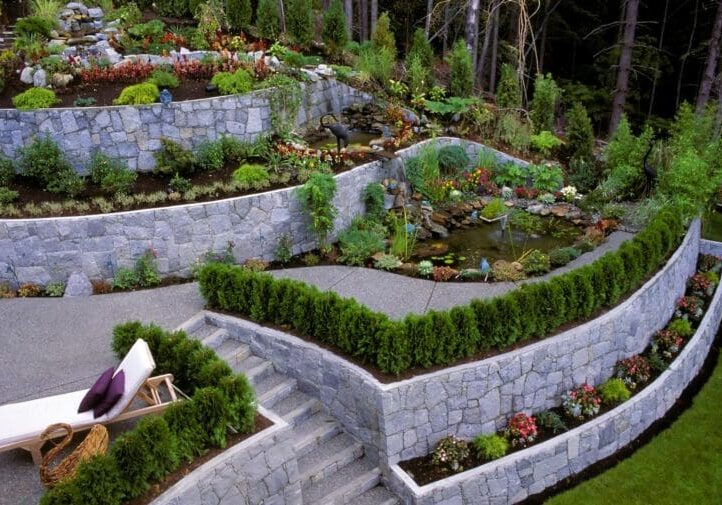 Multi-level terraced garden with stone retaining walls, pond water feature, lush plantings, and lounge seating on a sloped yard