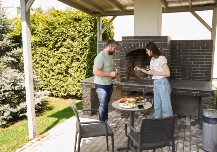Couple grilling at a built-in brick outdoor fireplace kitchen under a covered patio surrounded by lush hedges