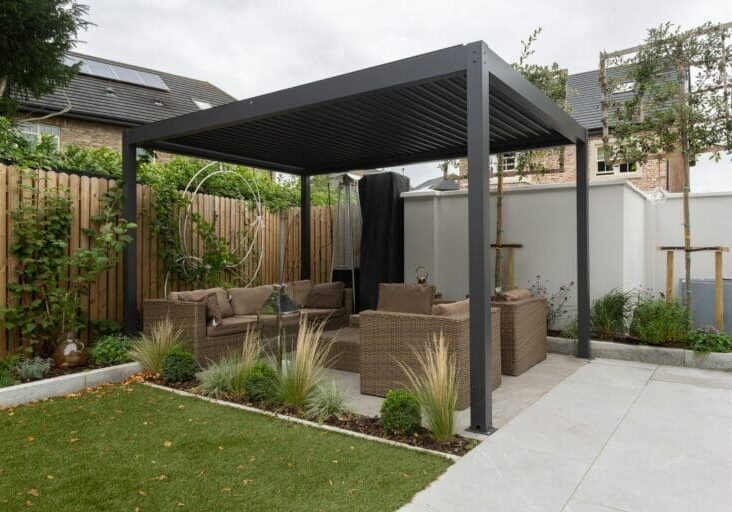 Modern dark aluminum pergola with wicker lounge seating, ornamental grasses, and stone paver patio in a backyard garden
