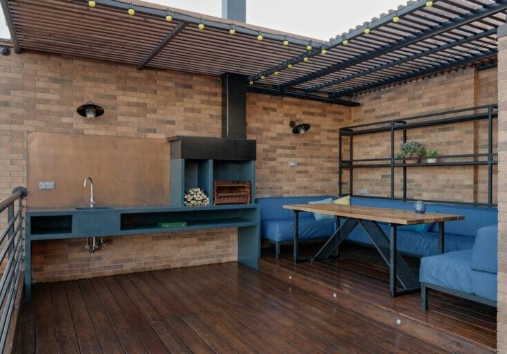 Rooftop outdoor kitchen with built-in wood-burning grill, sink, blue lounge seating, and pergola on a hardwood deck